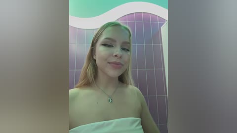 Video of a young Caucasian woman with long blonde hair, wearing a white towel, standing in a tiled bathroom with purple and teal accents.