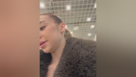 Media: Video of a close-up, side profile of a fair-skinned woman with straight, light brown hair tied back. She's wearing a black fur coat and has a slight, playful expression. Background features a grid-like, tiled ceiling.