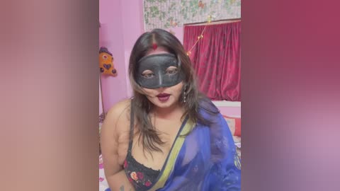 Media: A video of a South Asian woman in a black mask, red lipstick, and a blue sari, with a floral-patterned bed in a pink room with a red curtain.