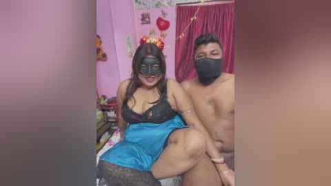 Media: Video of two people in a bedroom, a woman with long hair and a mask, wearing a black lace bra and blue satin skirt, sitting on a bed with a man wearing a mask and no shirt.