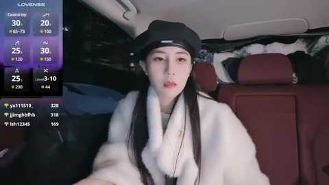 Media: Video of a young East Asian woman with long black hair, wearing a black beanie, white fur coat, and red lipstick, sitting in a car, with a digital display showing weather data.