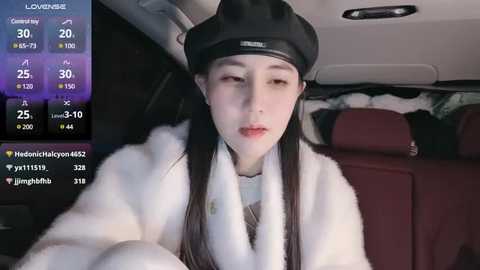 Media: Video of a young East Asian woman with fair skin, long black hair, wearing a black beret and white fur coat, sitting in a car with a plush red interior.