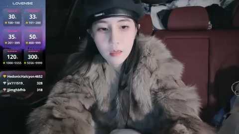 Media: Video of a young East Asian woman with pale skin, wearing a fur coat and black cap, sitting in a car. She has long, straight black hair.