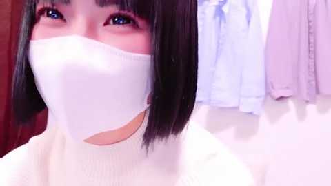 Media: Video of an East Asian woman with straight black hair and light skin, wearing a white face mask and a white top, standing in front of a pink and blue shirt hanging on a wall.