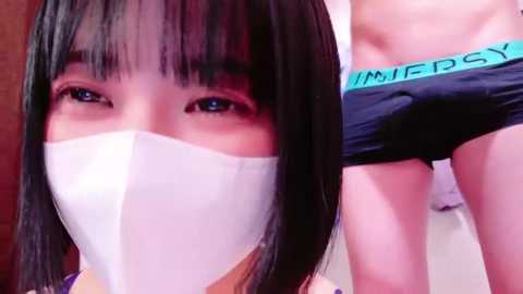 Video of an Asian woman with straight black hair, wearing a white surgical mask, and a man in black boxer shorts with \"IMPERIAL\" waistband.