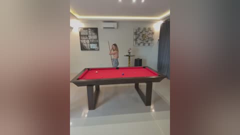 Media: Video of a woman with long hair in a white tank top and jeans playing pool in a modern, well-lit room with white walls, a red pool table, and minimalist decor.