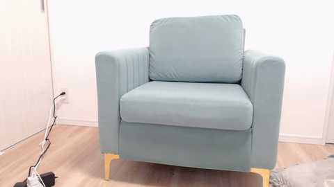 Media: A video of a pale blue, upholstered armchair with wooden legs placed on a light wooden floor in a minimalist room.