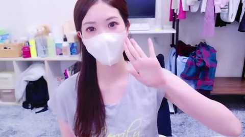 Media: Video of an Asian woman with long brown hair, wearing a white mask, gray shirt, and raising her hand in a room with a white desk, clothes hanging, and a soft gray carpet.