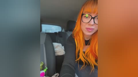 A video of a young woman with vibrant orange hair, wearing black-rimmed glasses and a black top, sitting in a car.