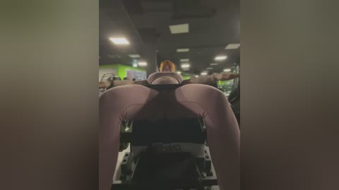 A video of a woman wearing tight pink leggings and a sports bra, bent over a stationary bike in a dimly lit gym, her legs prominently displayed.