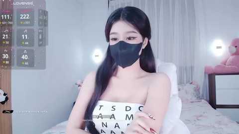 Media: Video of an East Asian woman with long black hair, wearing a black face mask and a white bandeau top with text, sitting in a brightly lit bedroom with a pink stuffed toy in the background.