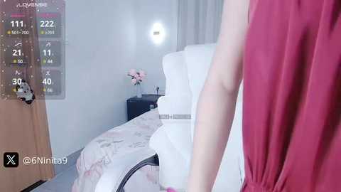 Media: A video of a woman in a pink sleeveless top, standing beside a white bed with a floral duvet in a minimalist, softly lit bedroom.