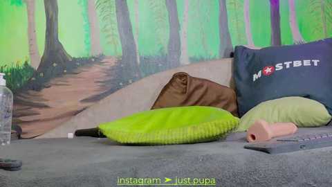 Media: Video of a cozy bed with a brown pillow and a lime green pillow, set against a green forest mural. \"MOSTBET\" pillow and a roll of paper towels on the grey bedspread.