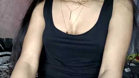 Media: A video of a woman with light skin and long black hair wearing a black sleeveless top, showing off her medium-sized breasts, necklaces, and a striped bed in the background.