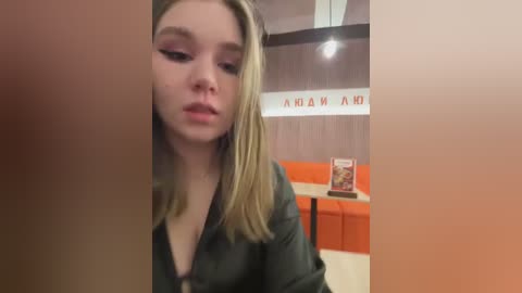 Video of a young Caucasian woman with straight blonde hair, wearing dark makeup and a black jacket, looking down, in a dimly lit restaurant with orange seats and \"AHA\" signage.