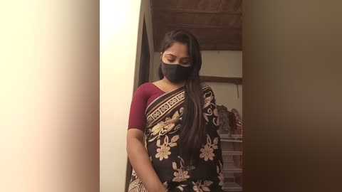 Media: Video of a South Asian woman with long black hair, wearing a maroon blouse and black sari with floral patterns, a black mask, and standing indoors with a soft, warm lighting.