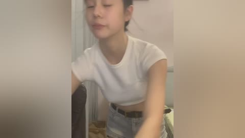 Media: Video of an East Asian woman with light skin and medium-length dark hair, wearing a white crop top and high-waisted jeans, leaning against a wall with a neutral background.