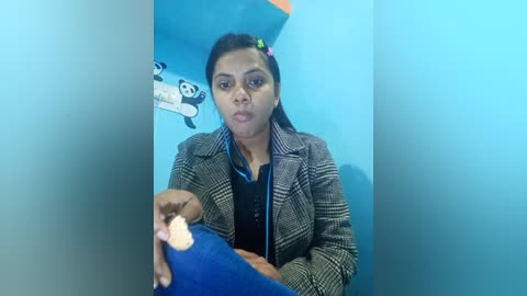 Media: Video of a South Asian woman with dark hair, wearing a plaid jacket over a blue shirt, holding a lit candle, in a blue-tiled bathroom with panda-themed stickers on the wall.