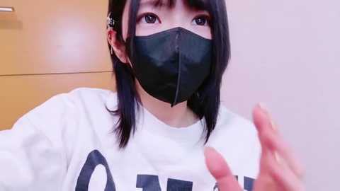 Media: Video of an Asian woman with short black hair, wearing a white sweatshirt with black text, black face mask, and gold earrings, making a peace sign. Background shows a light-colored wall and a wooden cabinet.