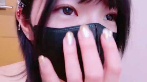 Media: Close-up video of an East Asian woman with long black hair, wearing a black face mask, brown eyes, and a silver earring, against a blurred beige and light brown background.