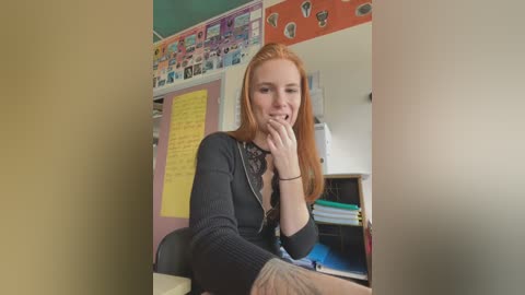 Media: Video of a smiling, red-haired woman in a black sweater, sitting at a desk in a colorful classroom. Background includes bulletin boards, papers, and educational posters.