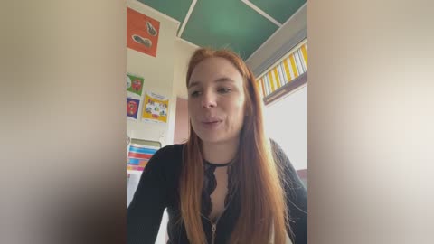 Media: Video of a fair-skinned, red-haired woman with long hair, wearing a black top, smiling in a colorful classroom with wall decorations and a window with yellow stripes.