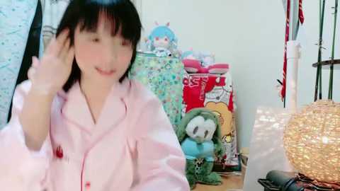 Media: Video of a young Asian woman with short black hair, wearing pink pajamas, talking on the phone in a cluttered, colorful room with toys, stuffed animals, and a red gift-wrapped box.