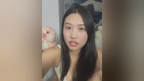 Media: Video of an Asian woman with long black hair, wearing a white tank top, taking a selfie in a dimly lit room. Background features a plush toy and a messy bed.