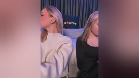 Media: Video of two blonde women in a studio, wearing white and black outfits, sitting side by side, with dark curtains and a black screen behind them.