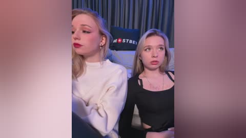 Media: Video of two women seated in a dimly-lit room, one with blonde hair and red lipstick, wearing a white sweater, and the other with blonde hair, wearing a black top, looking surprised.