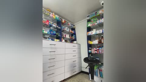 Media: Video of a small, well-lit pharmacy with white cabinets, shelves, and drawers filled with various medicines. A black scale is positioned near the center, with a white wall and light gray floor completing the scene.