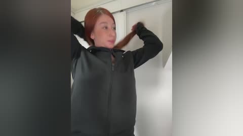 A video of a fair-skinned, red-haired woman in a black jacket, pulling her hair back with both hands. She stands in a room with white walls and a white shower curtain. The image has a blurred background.