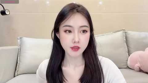 Media: A video of a young East Asian woman with long black hair, fair skin, and light makeup, sitting on a beige sofa. She wears a white top. Background includes a wall and a black light fixture.