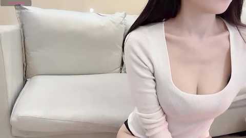 Video of a fair-skinned woman with long dark hair, wearing a tight white long-sleeve top, highlighting her cleavage, standing near a white couch in a minimalist living room.