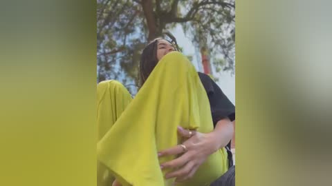 Media: A video of a woman in a bright yellow dress, sitting with her legs bent, surrounded by green foliage, with a blurred background.