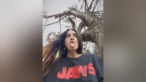 Media: Video of a young woman with long dark hair, wearing a black JAWS t-shirt, sticking her tongue out, in a forest setting with a large, twisted tree in the background.