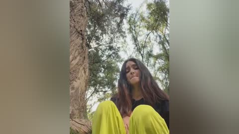 Media: Video of a young woman with long, dark hair, sitting in a forest. She wears a black top and bright yellow pants. Trees and greenery surround her, creating a serene outdoor setting.