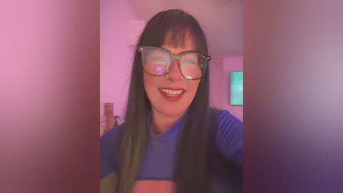 Media: Video of a smiling woman with long black hair, wearing large glasses and a blue crop top, in a room with pink and purple lighting.