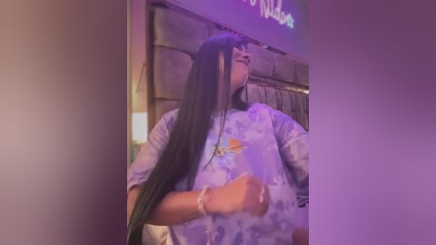 Media: Video of a woman with long black hair, wearing a floral-patterned shirt, sitting in a dimly lit room with purple lighting.