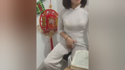 Media: A video of an Asian woman in traditional white Chinese dress, seated in a dimly lit room, holding a book, with decorative red lanterns and green ribbons on the wall.