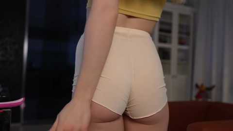 Media: Video of a woman's lower back and buttocks in tight, cream-colored shorts, wearing a yellow crop top, in a dimly lit, modern living room with glass shelves and a pink toy in the background.