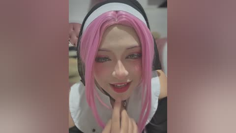 Media: Video of a young, fair-skinned woman with pink hair and light makeup, dressed as a nun, kneeling, with a male hand holding her chin. Background is blurred.