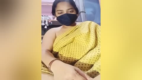 Media: Video of a woman with medium brown skin, wearing a black mask, a yellow polka-dot sari, and a brown wristband, sitting in a dimly lit room with blurred background.