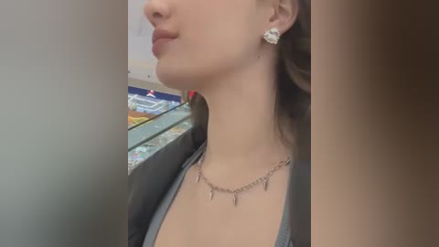 Media: Video of a woman with fair skin, wearing a black cardigan over a gray top, and a silver necklace with small tassel pendants. She has light brown hair and is seen from the side in a store aisle with colorful products in the background.