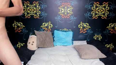 Media: A video of a dark room with ornate, colorful wallpaper featuring floral patterns. A naked man is partially visible on the left, standing near a bed with white sheets and two pillows, one light gray, one turquoise.