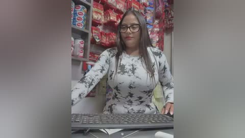 Media: Video of a middle-aged Latina woman with long dark hair, glasses, and a white tie-dye top, sitting at a desk in a convenience store, surrounded by colorful snack shelves.