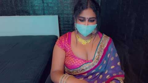 Media: Video of a South Asian woman in a colorful saree, wearing a face mask, sitting on a bed. She has medium brown skin, long dark hair, and is adorned with gold jewelry.