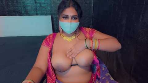 Media: A video of a South Asian woman in a pink sari, partially exposing her large breasts with a hand, wearing a blue mask and gold jewelry. She is seated on a bed with dark walls.