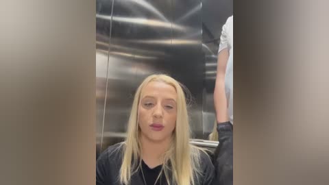 Media: Video of a young blonde woman with light skin, wearing a black jacket, seated in a stainless steel elevator, surrounded by modern, industrial architecture.