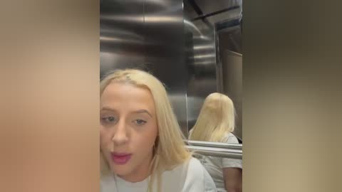 Media: Video of a blonde woman in a white shirt, with a surprised expression, inside a metallic elevator, looking at her reflection in the mirrored wall.